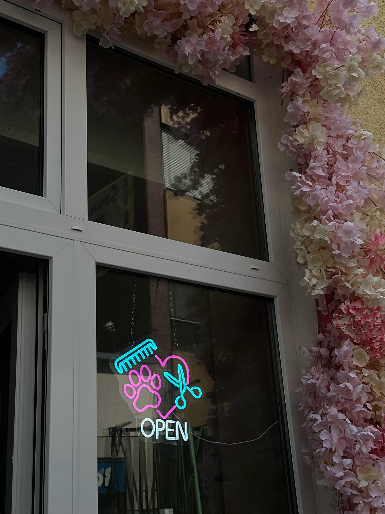 Schaufensterfront des Pfotenparadies mit rosa Blütenbogen und Neon-OPEN-Schild in Form von Schere, Kamm und Pfote